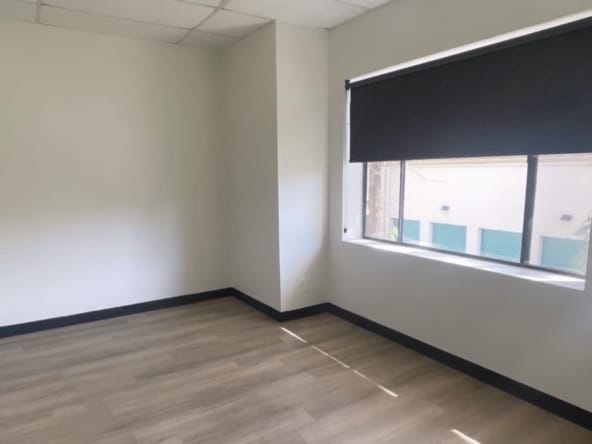 Bright empty salon room with large window, black blinds, and wood flooring for lease.