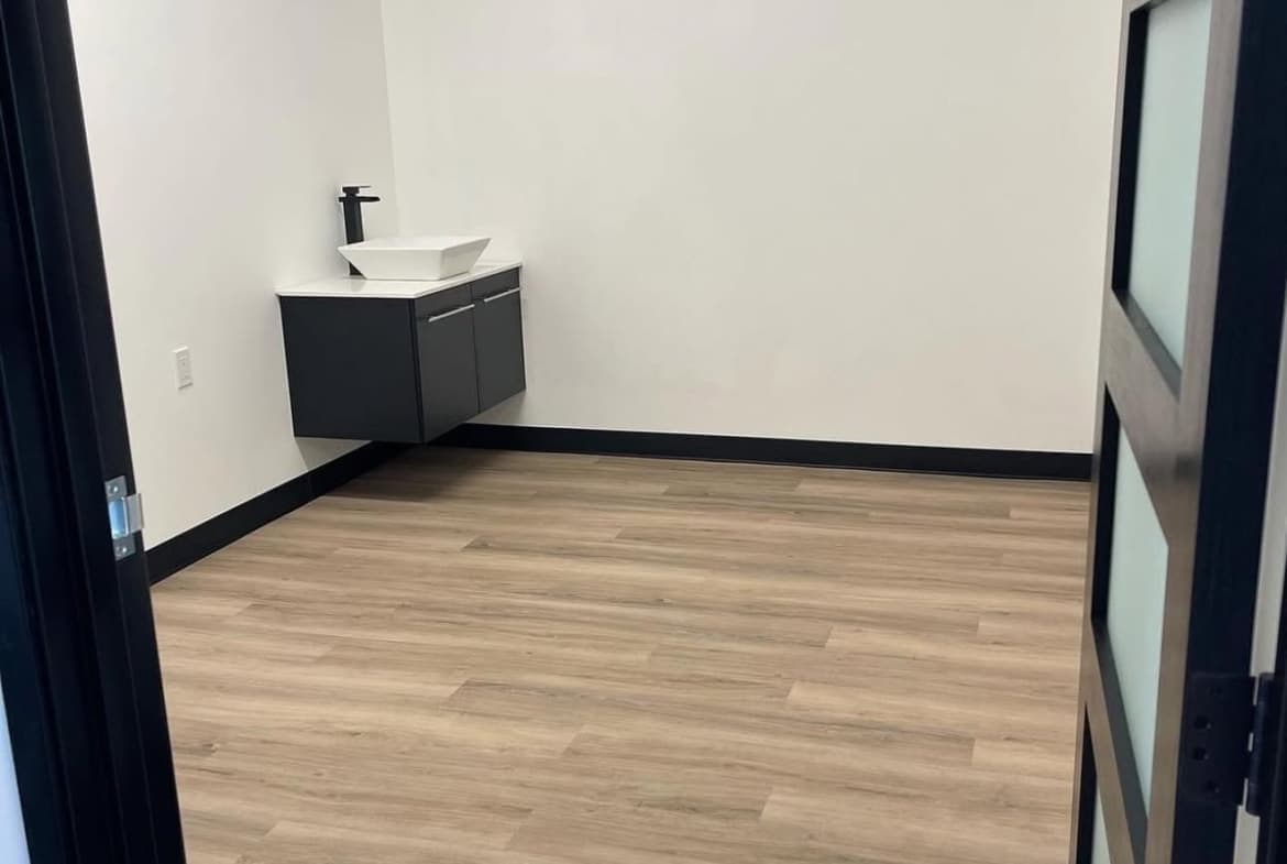 A clean, minimalist salon room featuring wooden floors, a sink, and a white wall, perfect for beauty business rental.