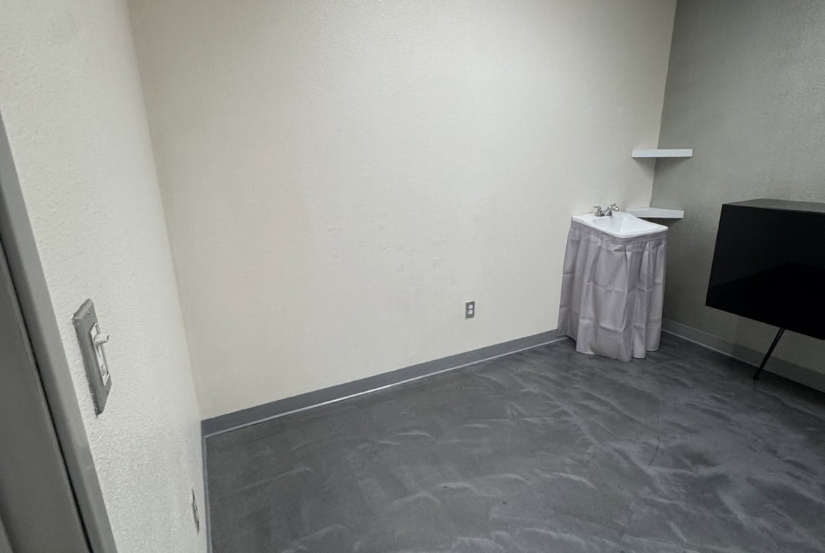 Empty salon rental room with gray flooring, white walls, and ceiling light for beauty professionals.