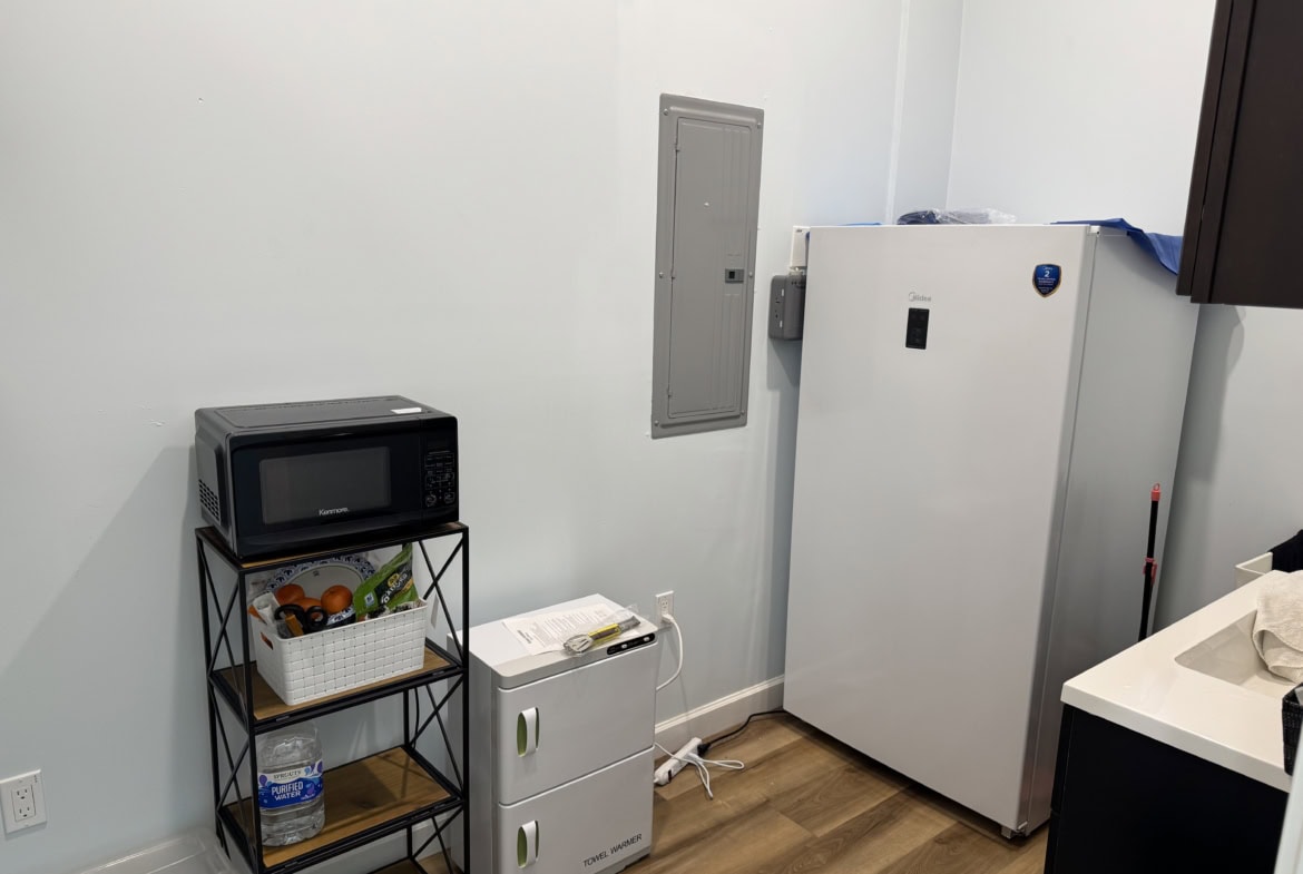 Utility room with refrigerator, microwave, and storage shelves at Salon Renter.