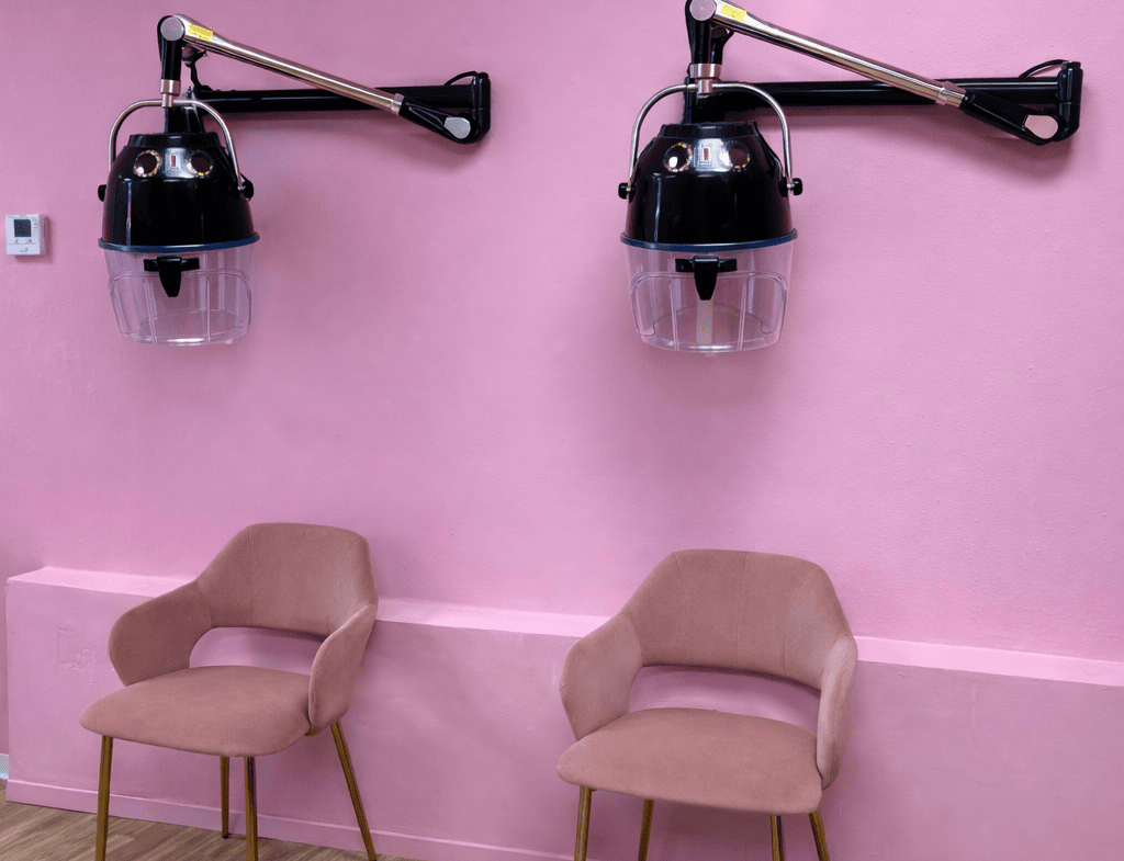 Two salon hair dryers mounted on a pink wall with two beige chairs below.