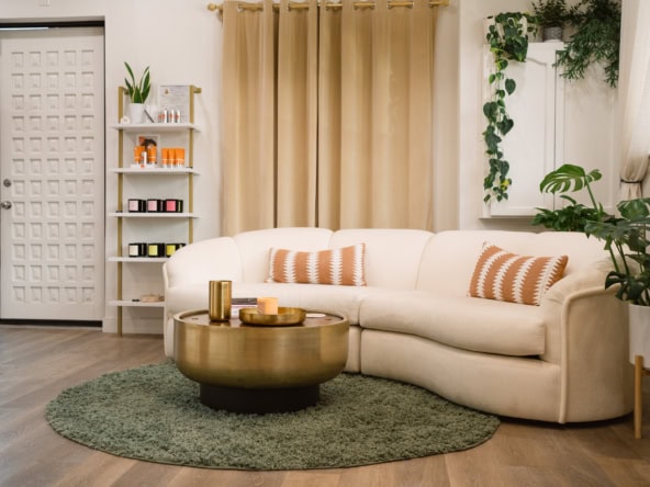 Modern salon waiting area with comfortable cream sofa, decorative pillows, and lush green plants. Pe.